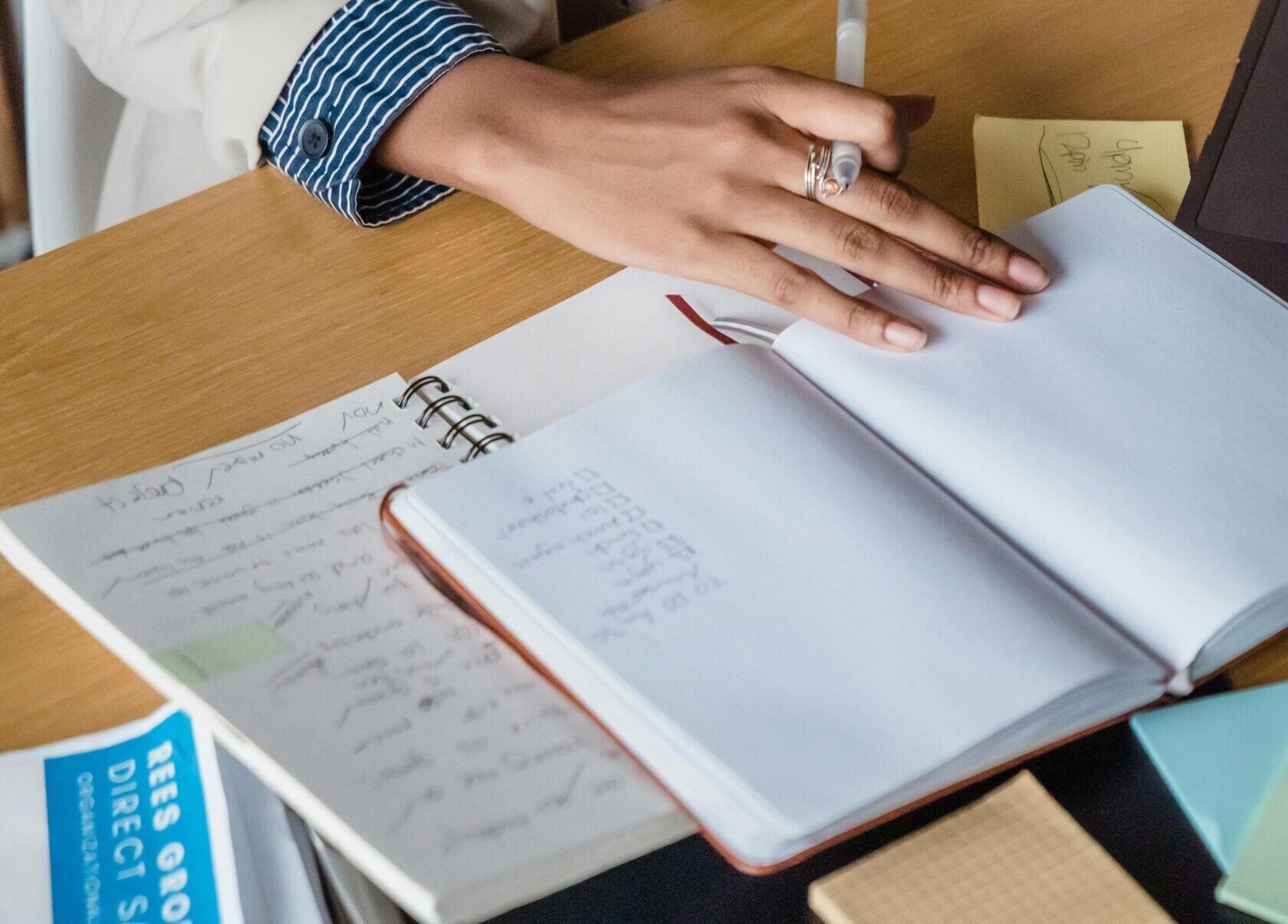 Woman writing in a notebook at a desk with papers and sticky notes, highlighting office productivity.